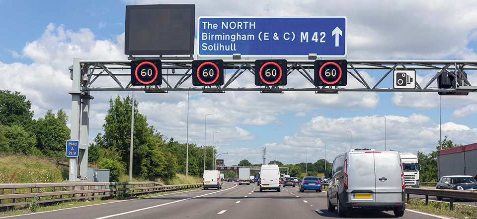 Motorway gantry showing four amber 60 mph roundels above lanes with a blue overhead direction sign for The North, Birmingham and Solihull on the M42